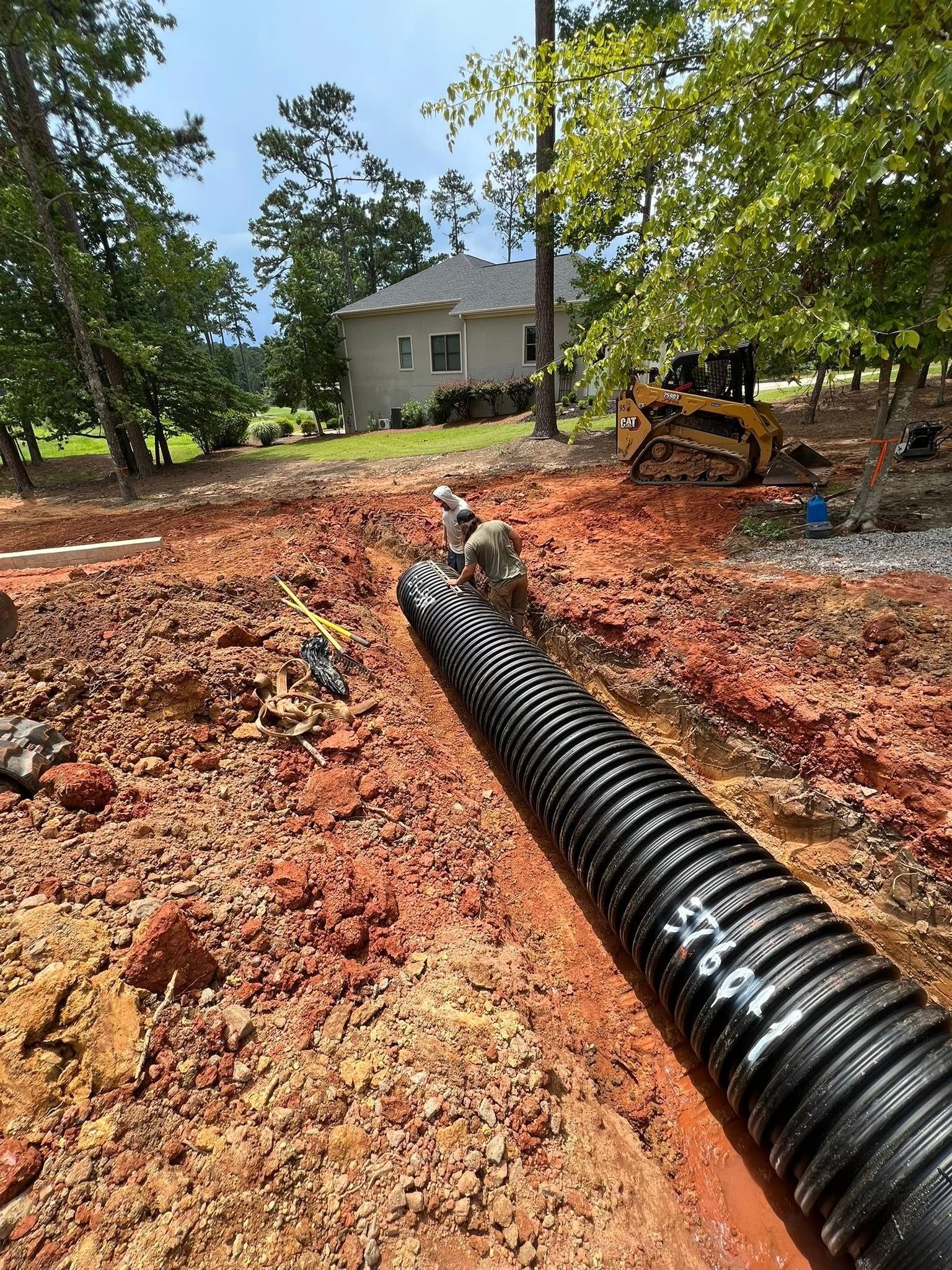 Black corrugated drainage pipe laid in a red dirt trench on a rural construction site near a house.