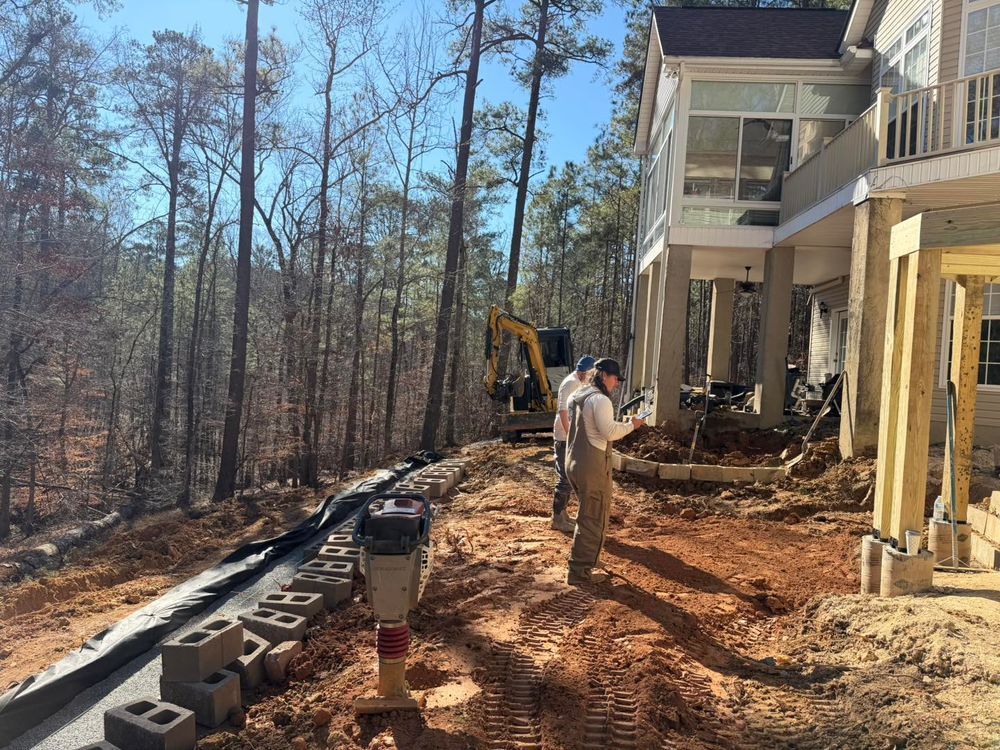 Construction workers grading dirt beside a beige house on a wooded slope, with cinder blocks and an excavator nearby.