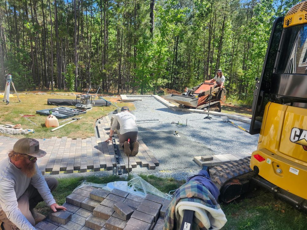 Workers laying gray pavers beside a yellow excavator in a wooded outdoor area