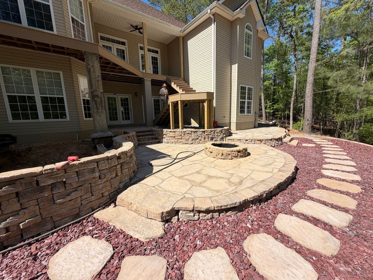 Curved stone patio with fire pit beside a house, bordered by red mulch and stepping stones in a wooded backyard