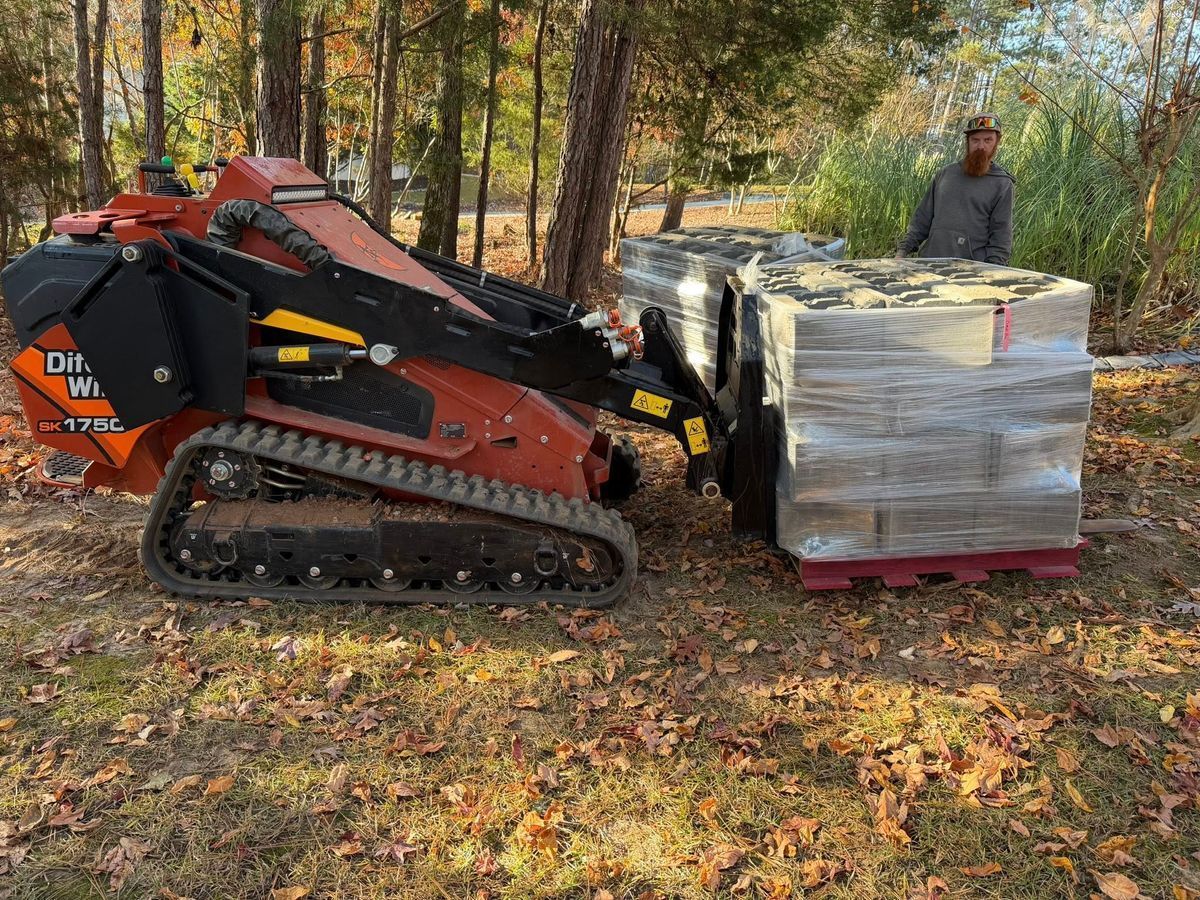 Orange tracked loader pushing a pallet of wrapped blocks in a wooded area, with a worker nearby