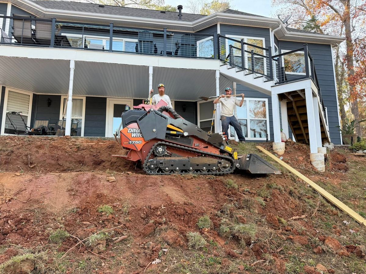 Worker on an orange excavator stabilizing a steep dirt slope beside a damaged house