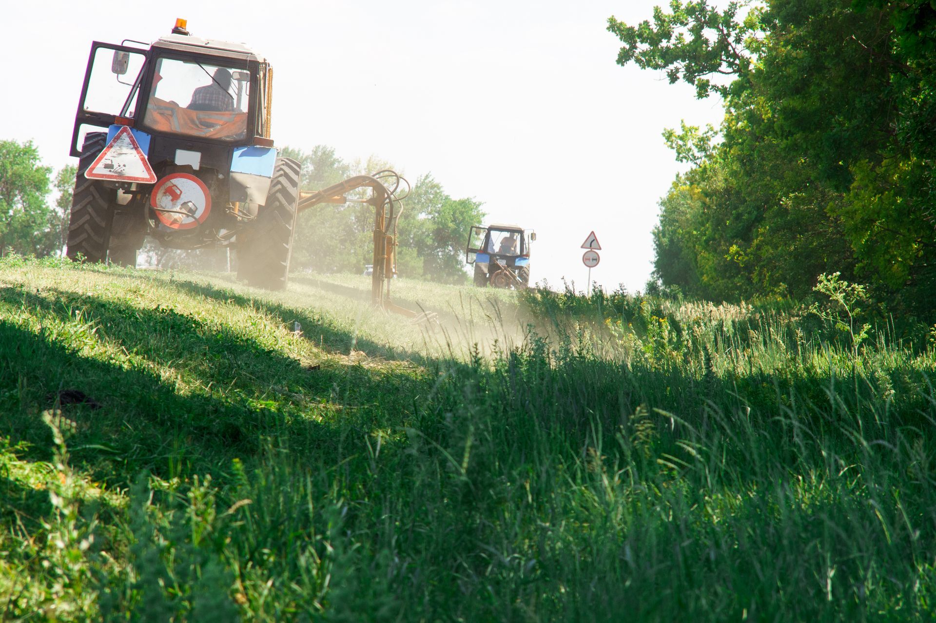 Tractors spraying a dusty green field along a sunny rural path, with trees in the background