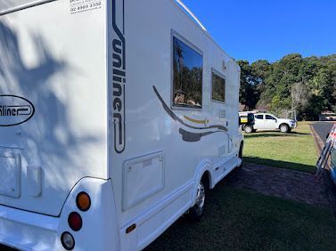White travel caravan parked on grass beside a road, with windows and a wooded background
