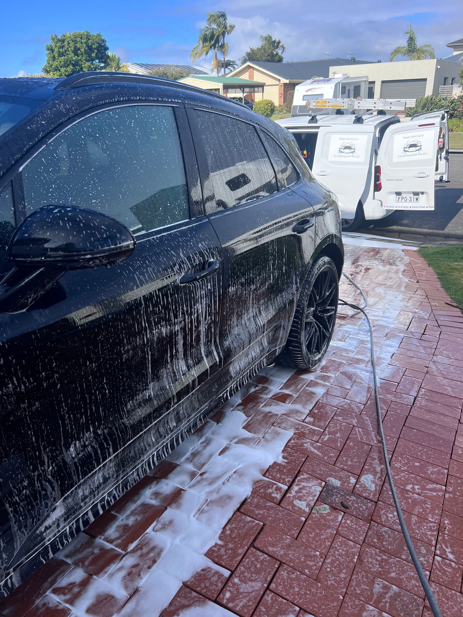 Black Car Being Washed With SOAP Foam In A Driveway, With Houses And Bright Sky In The Background — Car Detailing Mobile Cleaning Co In Thrumster, NSW
