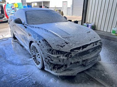 Grey sedan covered in thick foam during a car wash, parked outdoors on wet pavement.