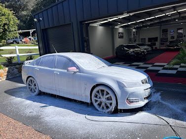 White sedan covered in foam at a car wash bay, with a garage and another car in the background