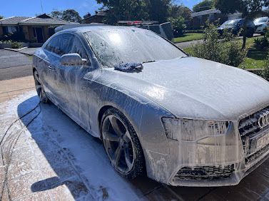 White Audi sedan covered in foam during an outdoor car wash on a sunny driveway