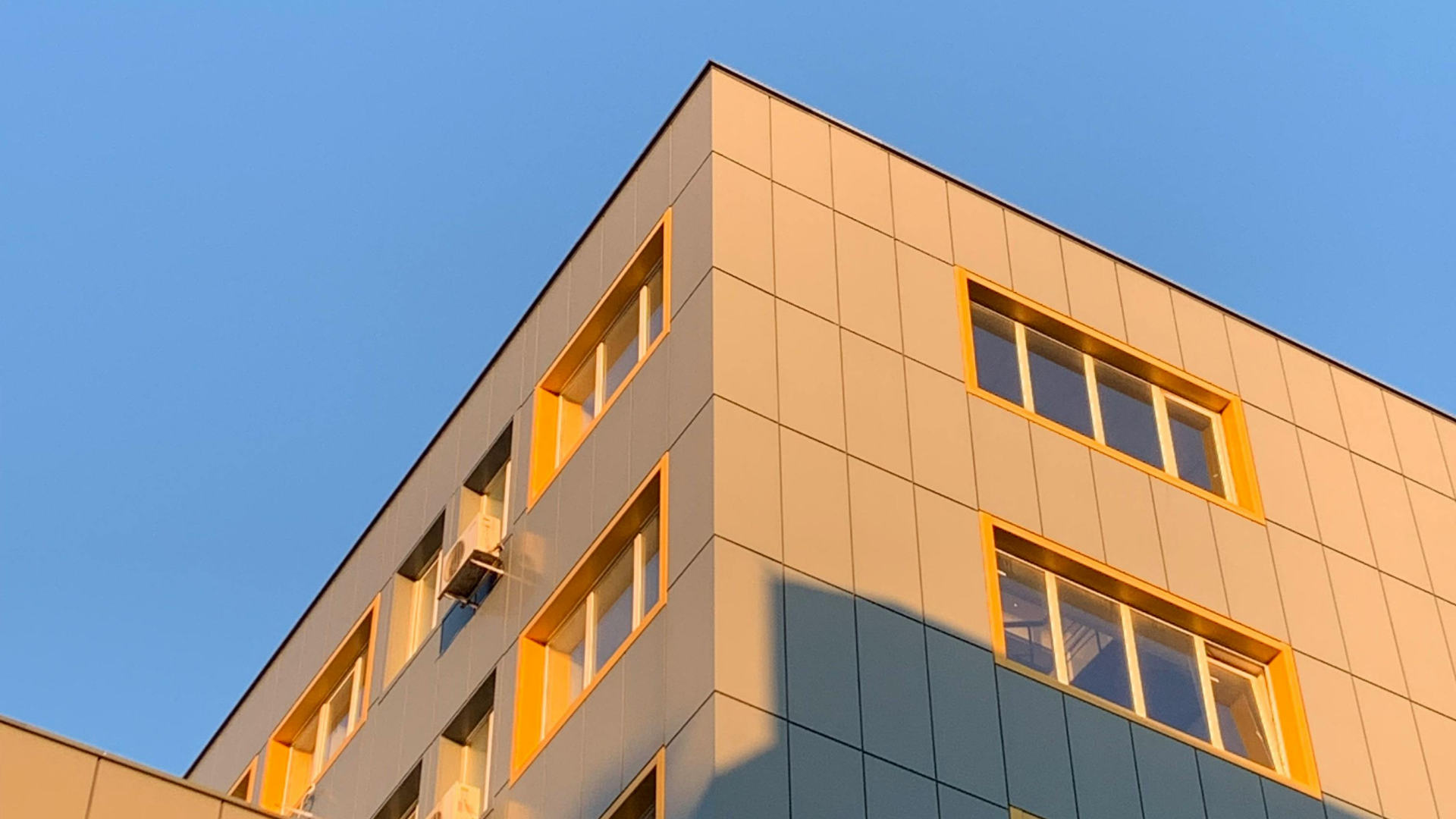 A low-angle view of a modern building facade with gray panels and yellow window frames against a clear blue sky.