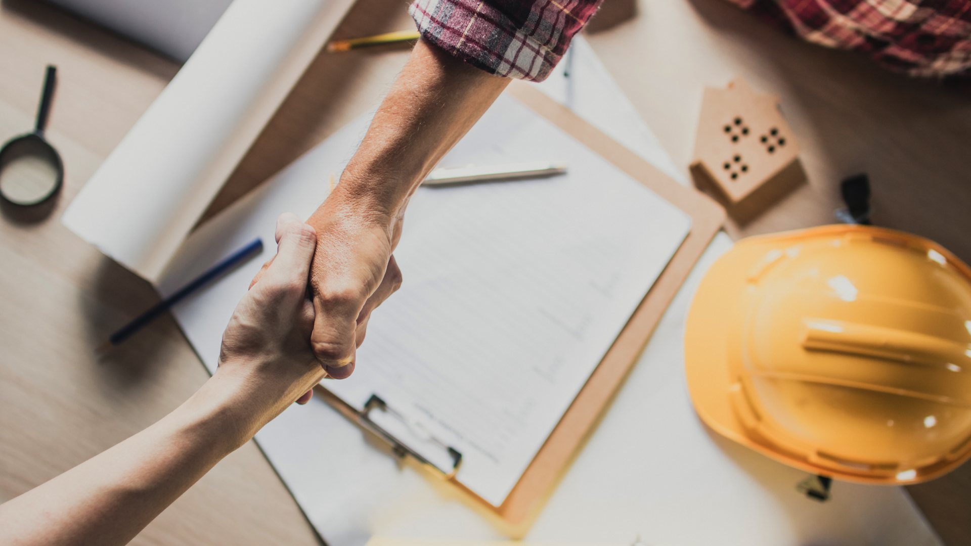 Two people shake hands over construction blueprints, a clipboard, and a yellow hard hat on a desk.
