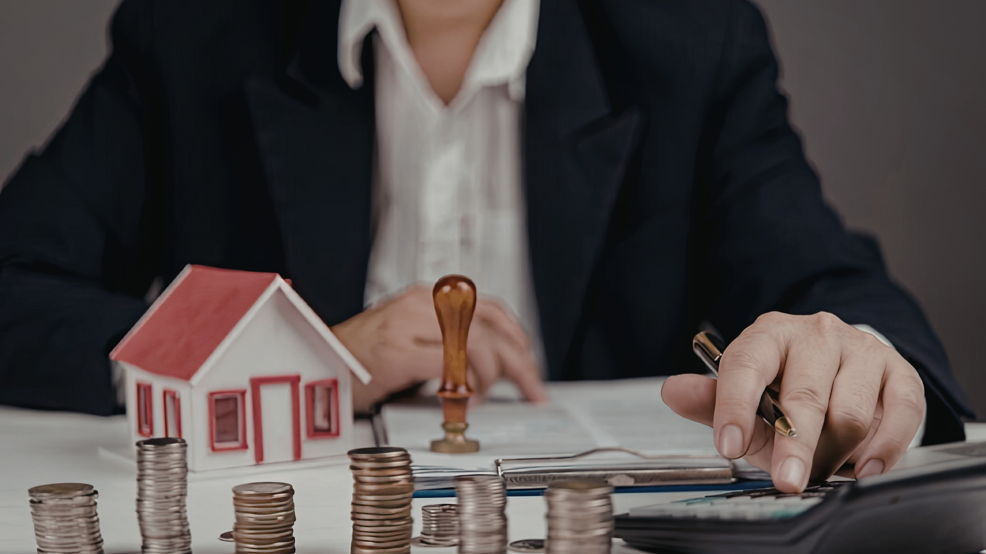 A person in a business suit works at a desk with a model house, stacks of coins, a document, and a calculator.
