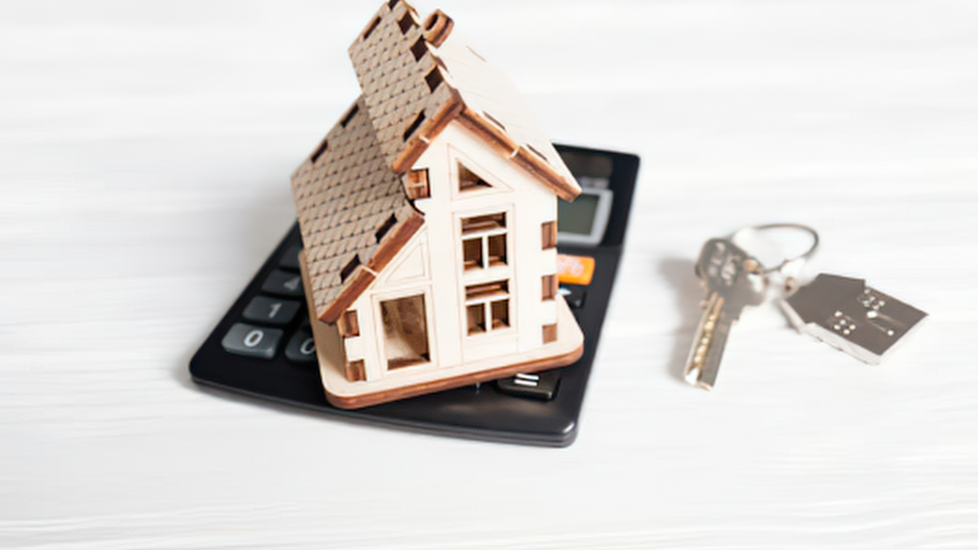 A small wooden house model sits on top of a black calculator next to a metal house key on a white surface.
