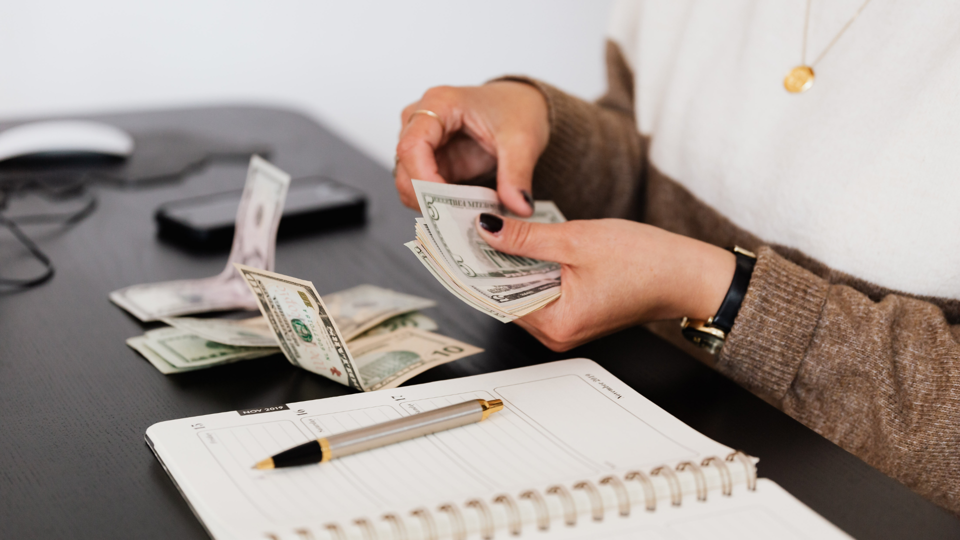 A person counting US dollar bills at a desk next to an open notebook and a pen.