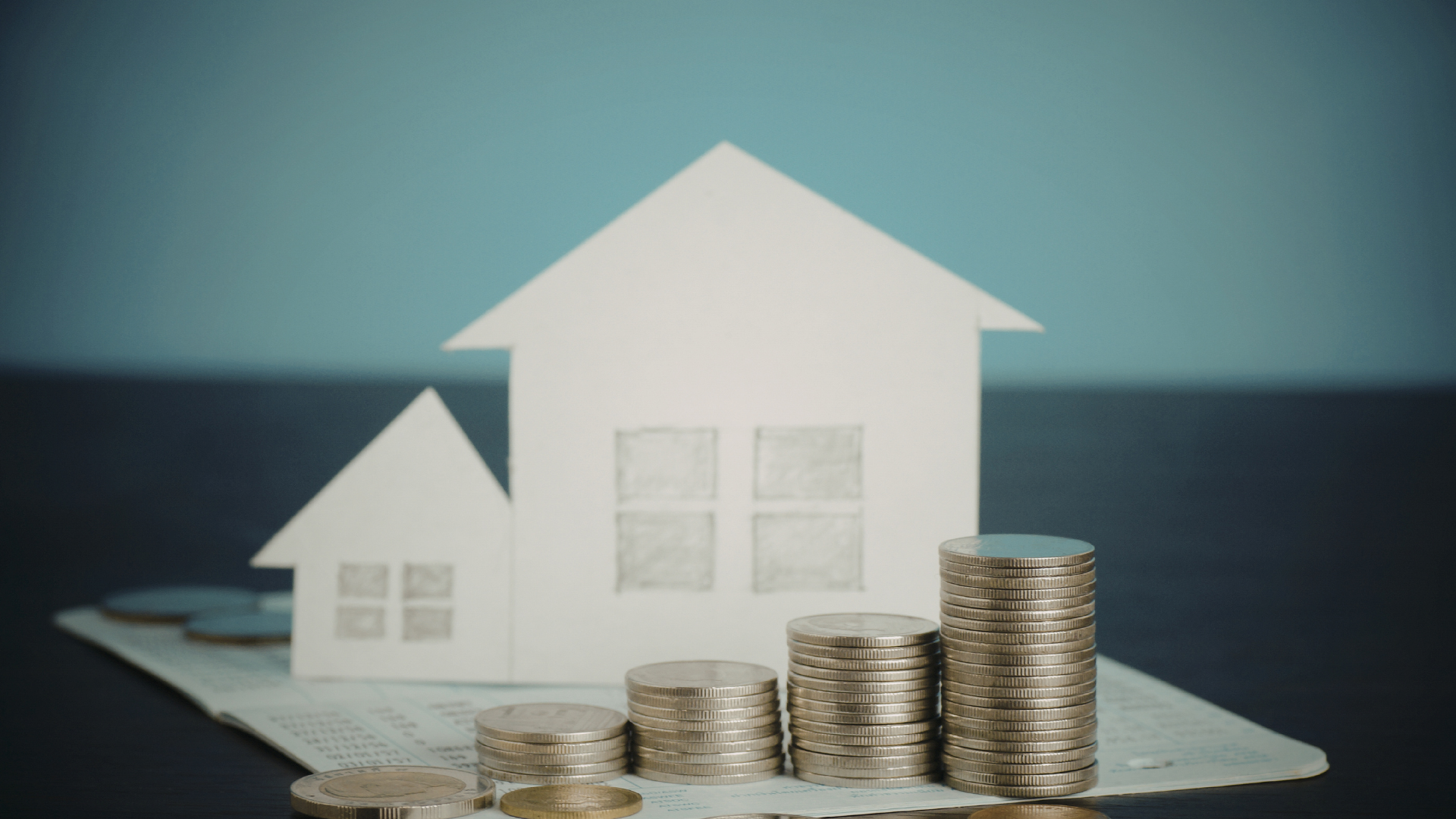 Two white model houses sit behind stacks of silver coins of varying heights, arranged on a flat surface.