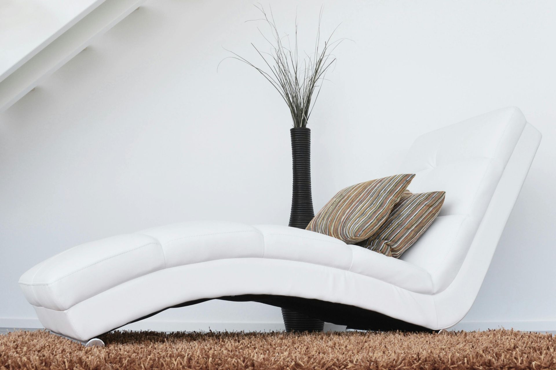 White chaise lounge with two pillows and a tall, dark vase with decorative branches against a white wall.