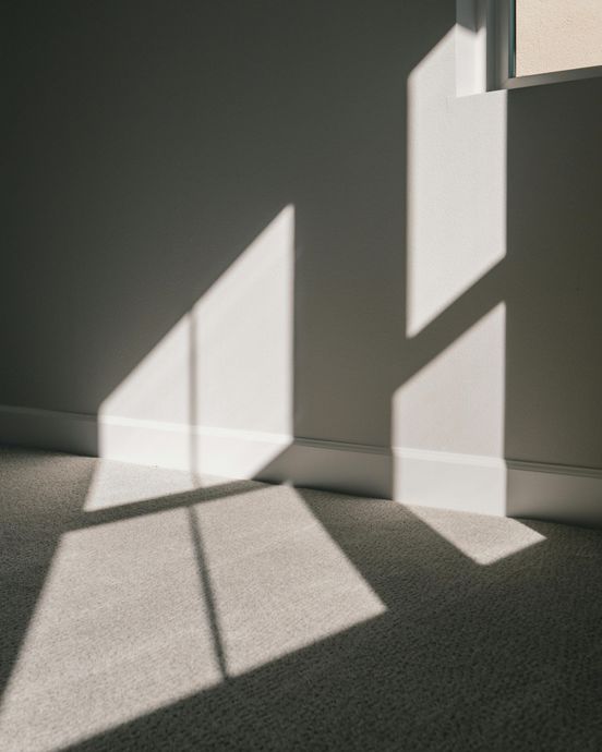 Shadows of a window frame on a white wall and carpet, creating geometric shapes with light.