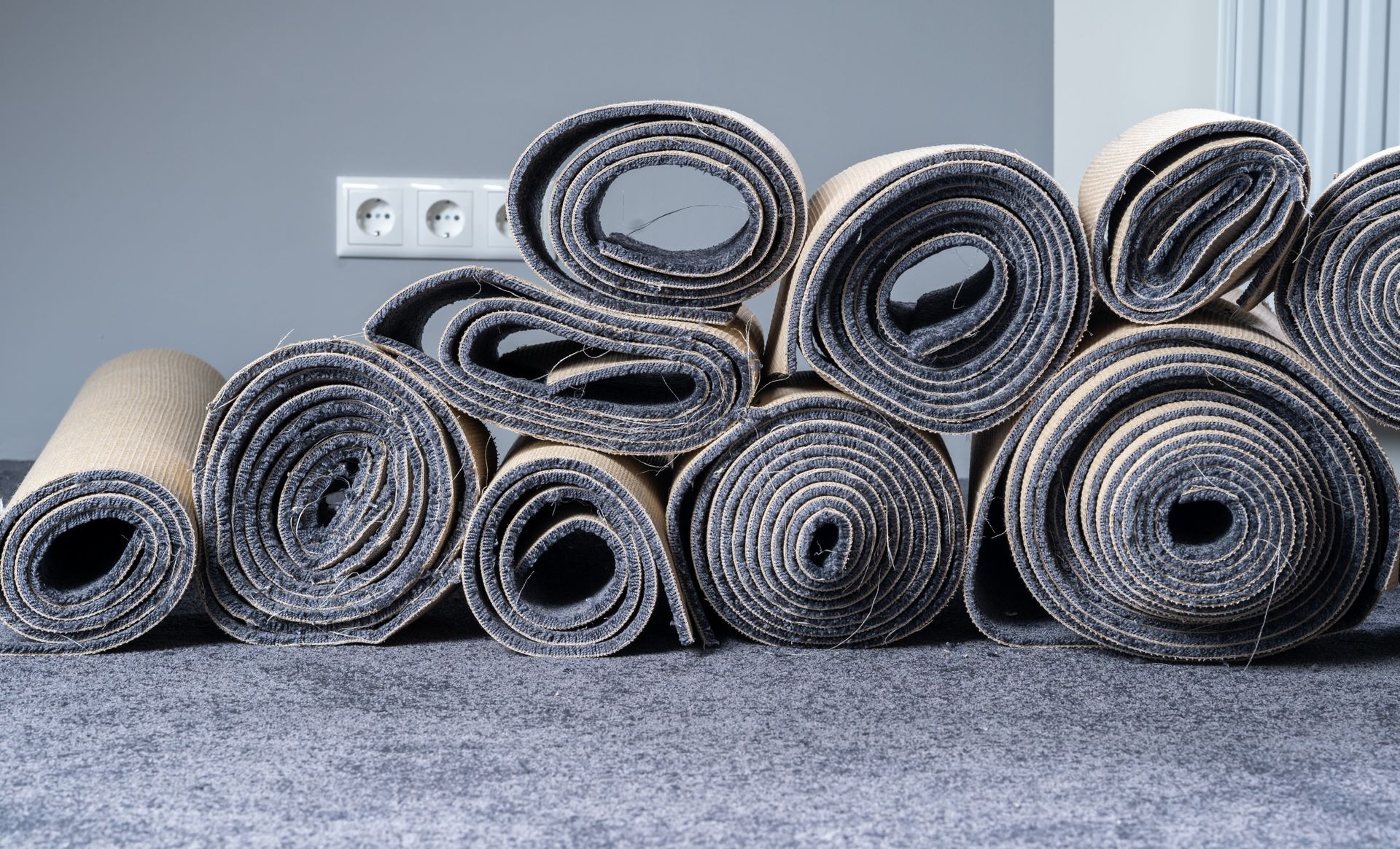 Rolled-up carpets of various sizes stacked against a gray wall, displayed on a larger carpet.