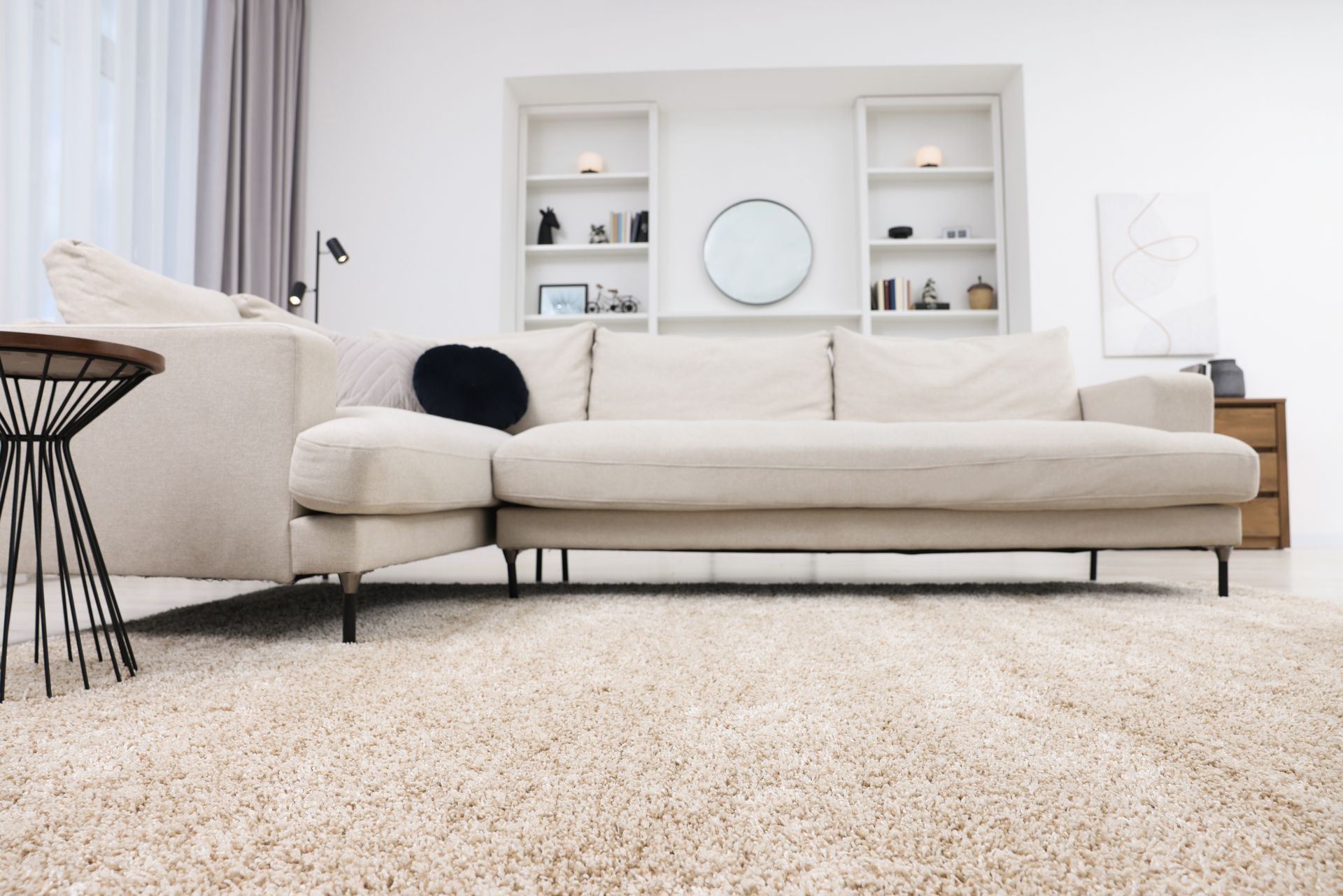 Beige sectional sofa in a bright living room, with shelves, round mirror, and shaggy rug.
