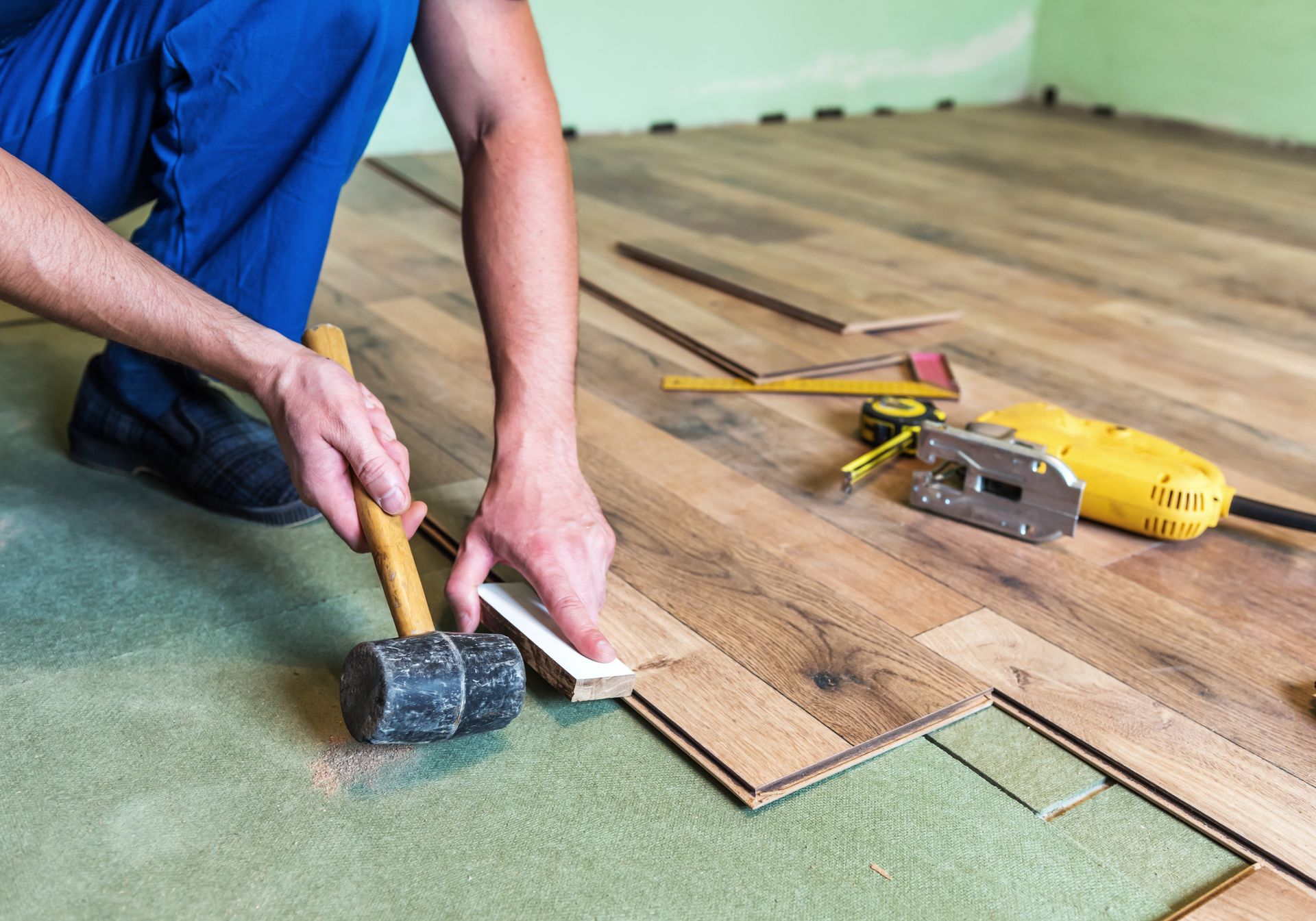 Person installing wooden floorboards, using a mallet and a white spacer in a room under construction.