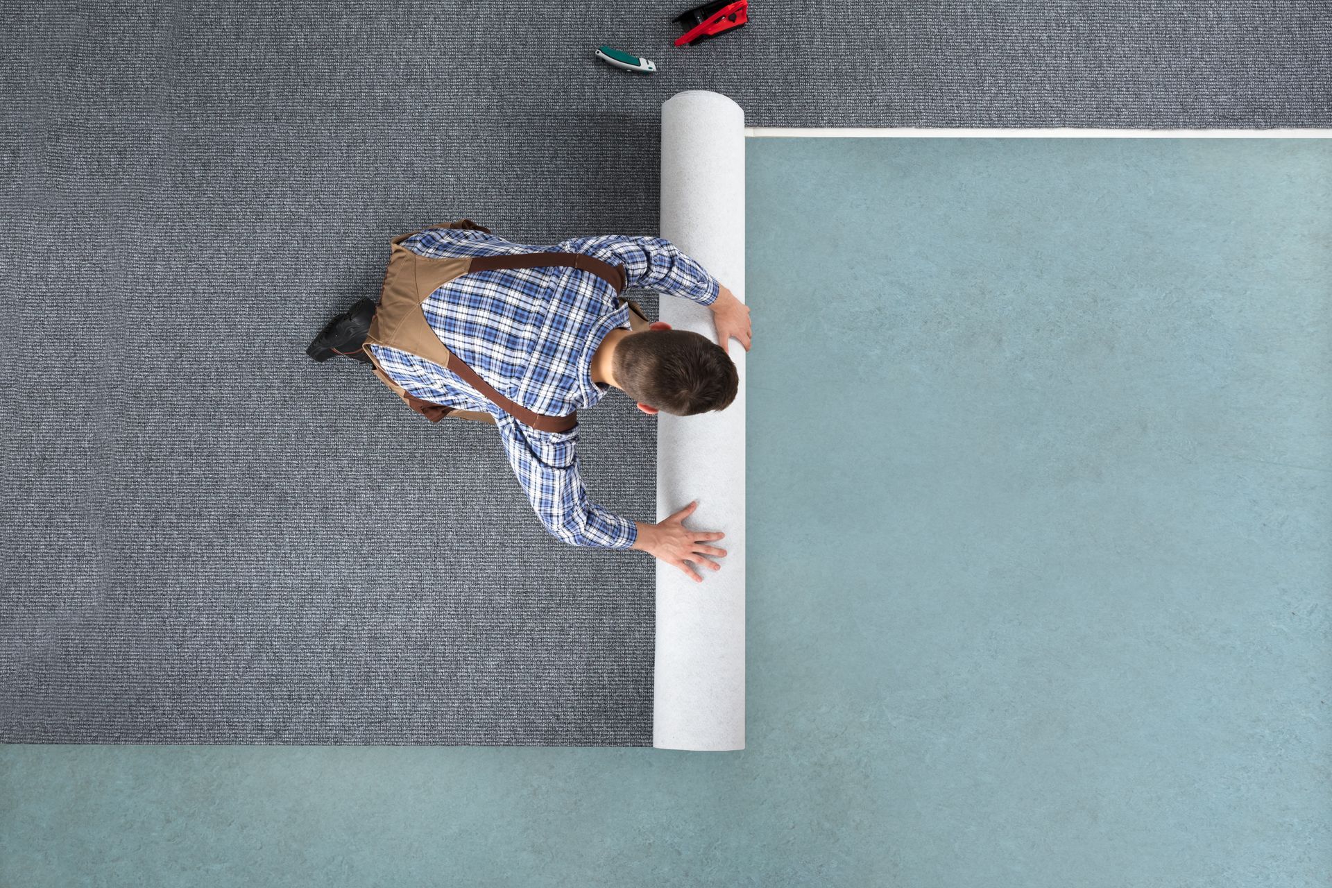 Person rolling out gray carpet onto a blue floor. Tools are placed above.