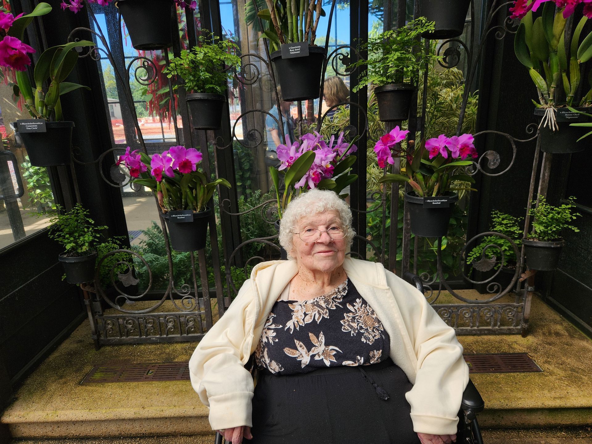 An elderly woman in a wheelchair is sitting in front of a wall of flowers.