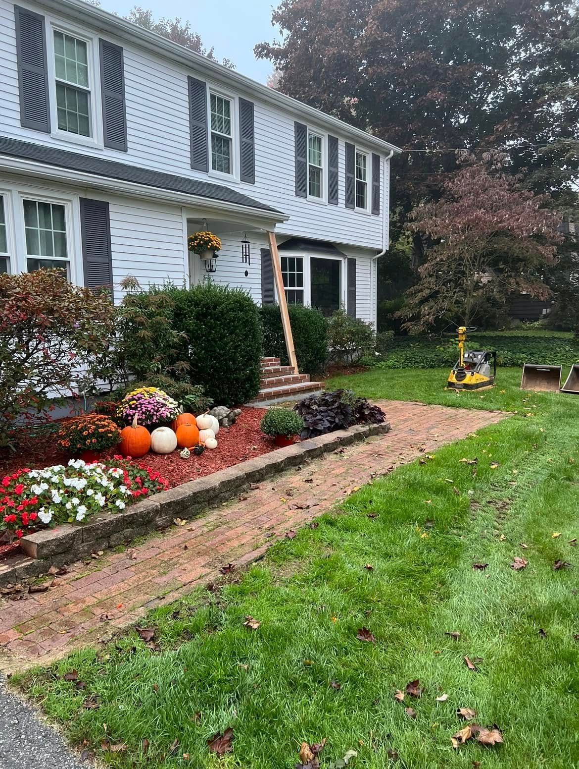 A two-story white house with a brick walkway and front yard landscaping featuring colorful fall flowers and pumpkins.