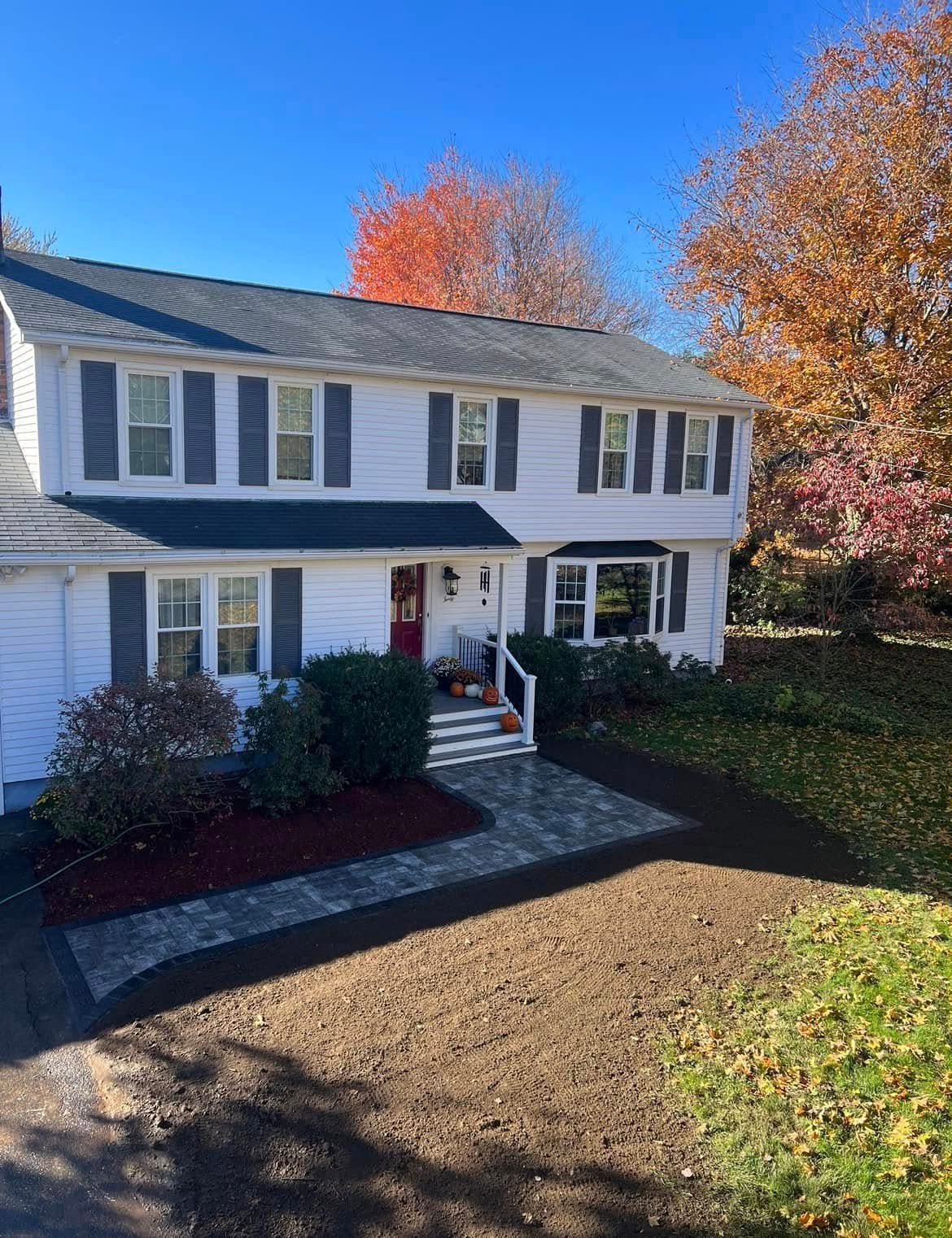 A two-story white house with a brick walkway and front yard landscaping featuring colorful fall flowers and pumpkins.