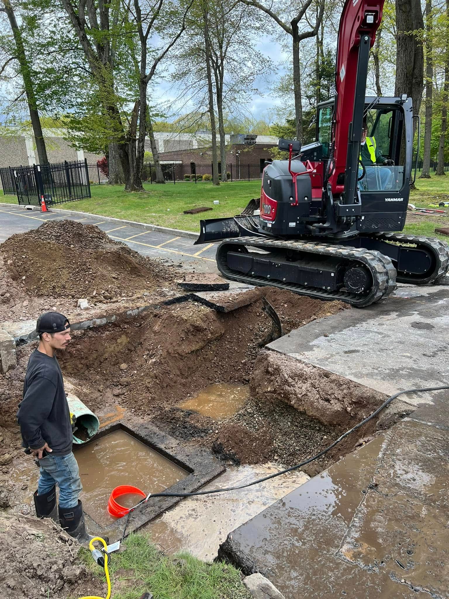 A person in a work shirt and jeans stands by a dirt trench next to a red excavator parked on a paved path.