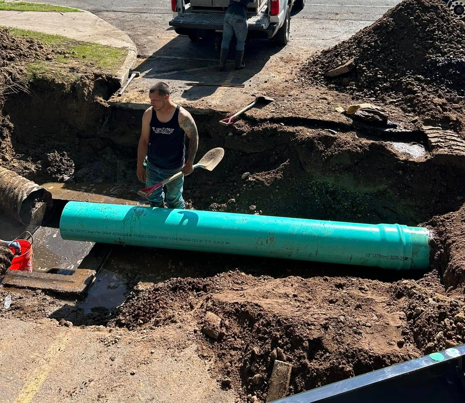 A worker stands in a trench next to a large green pipe being installed underground.