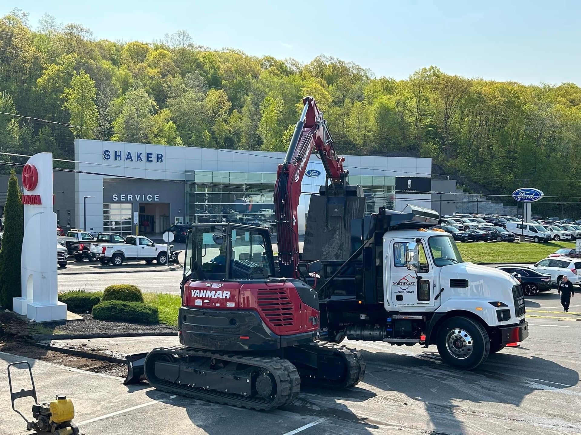A red tracked excavator loading debris into a white dump truck in front of a Toyota dealership service building. A red tracked excavator loading debris into a white dump truck in front of a Toyota dealership service building.