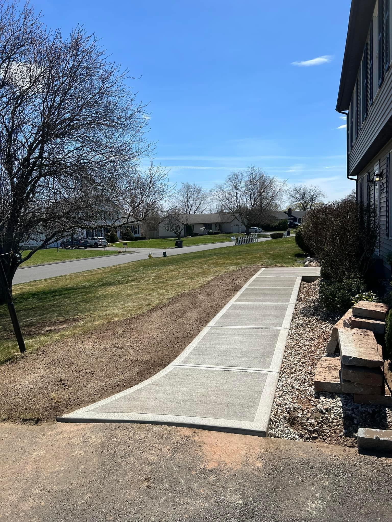 A concrete walkway leads from a driveway toward the side of a suburban house, flanked by a grassy lawn and landscaping rocks.