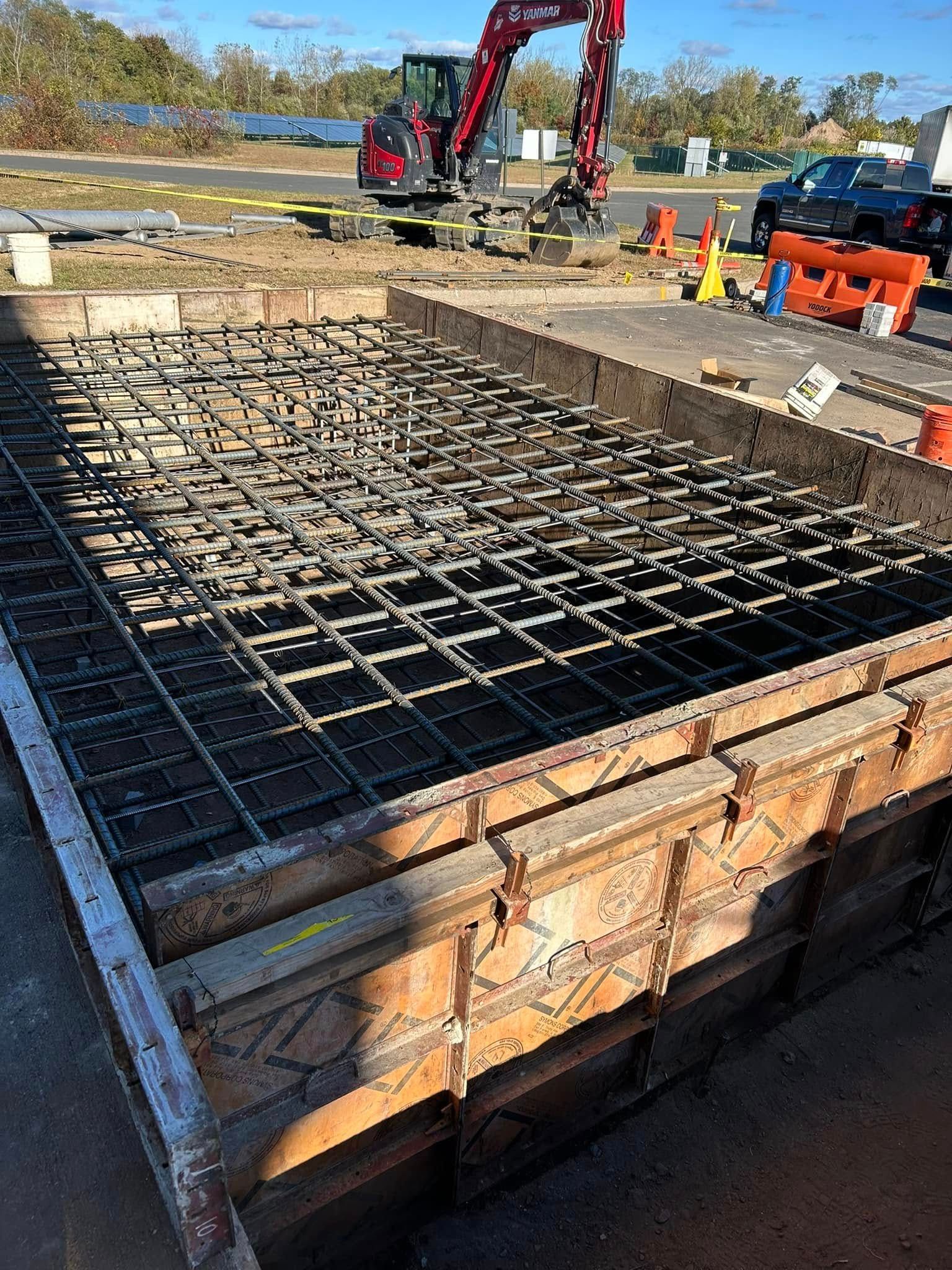 A reinforced concrete foundation form filled with a metal rebar grid at a construction site with an excavator nearby.