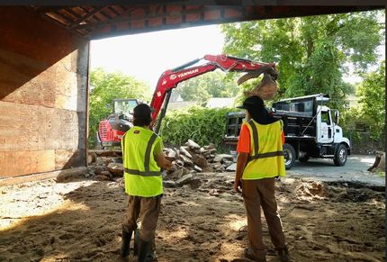 Two workers in bright safety vests watch a red excavator loading rubble into a white dump truck at a construction site.