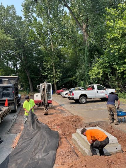 Workers in high-visibility gear install a concrete structure in a roadside excavation with a tractor and pickup truck nearby.