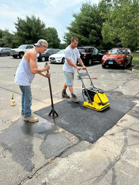A red excavator sits on a paved path next to a dirt excavation site, with a worker nearby at a construction project.