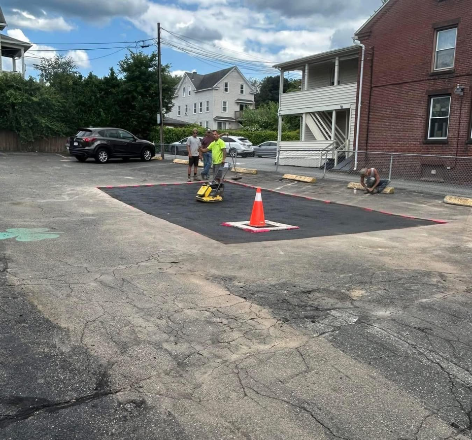 Two people in high-visibility vests work on a freshly paved patch of asphalt in an outdoor parking lot.