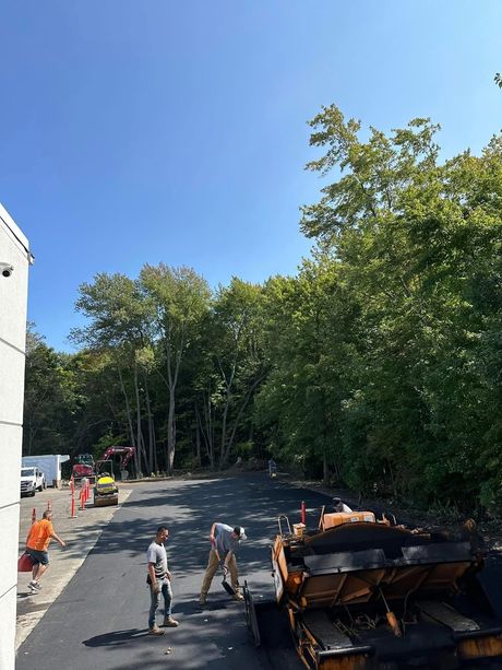 Workers pave a parking lot near a tree line on a sunny day, with a construction vehicle in the foreground.