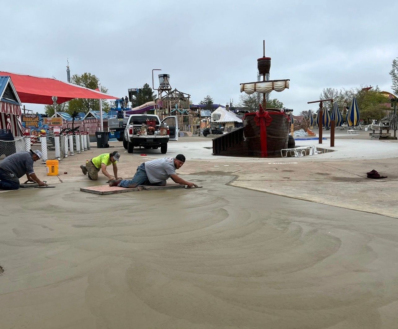A small excavator works on a backyard pool demolition project near a large red dumpster and dense forest trees.