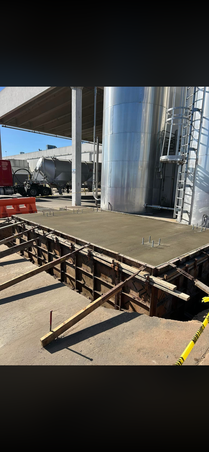 A person stands beside a child operating an orange paving machine on a gravel path near a wooded area.