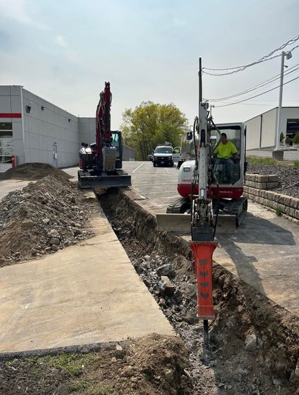 A worker guides a concrete structure lowered by a Takeuchi excavator into a trench at a residential construction site.