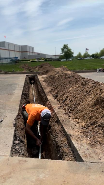Two construction workers install a concrete storm drain basin into the ground next to a small excavator and a truck.