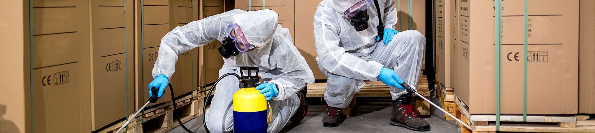 Two people in hazmat suits spray for pests in a warehouse.