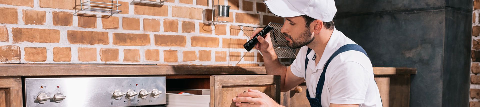 A chef in a kitchen, using a flashlight to examine a wooden cabinet. Brick wall in background.