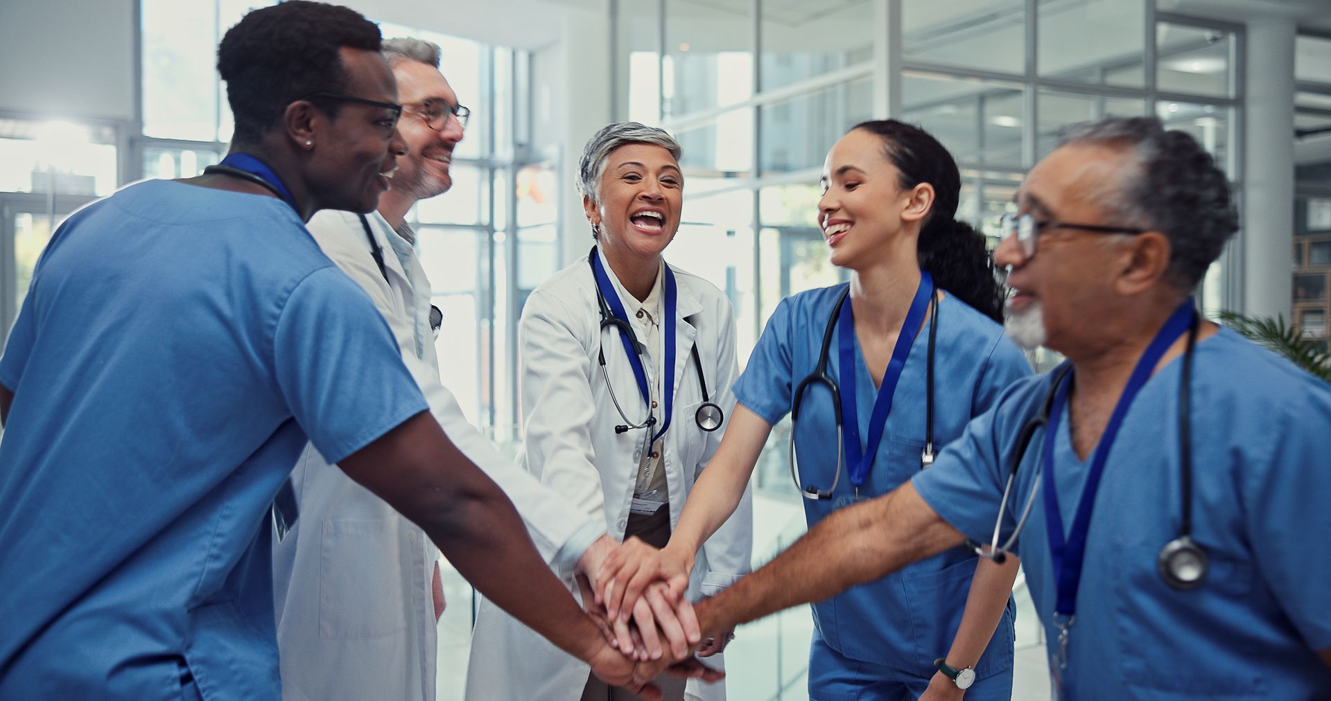 Medical staff in scrubs and white coats standing in a circle with their hands stacked in a gesture of unity.