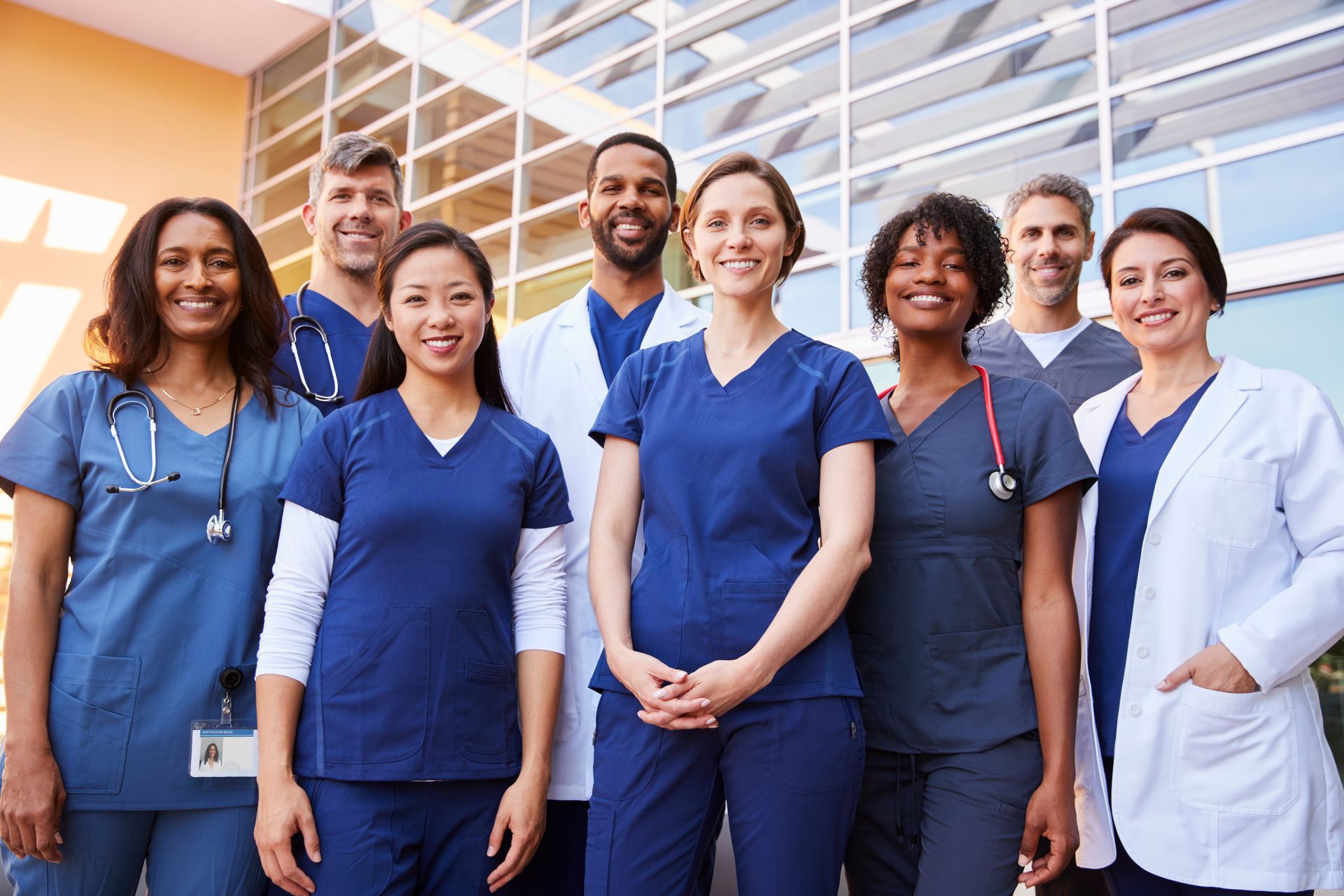 Medical professionals smiling in front of a modern building; many wearing scrubs or white coats.