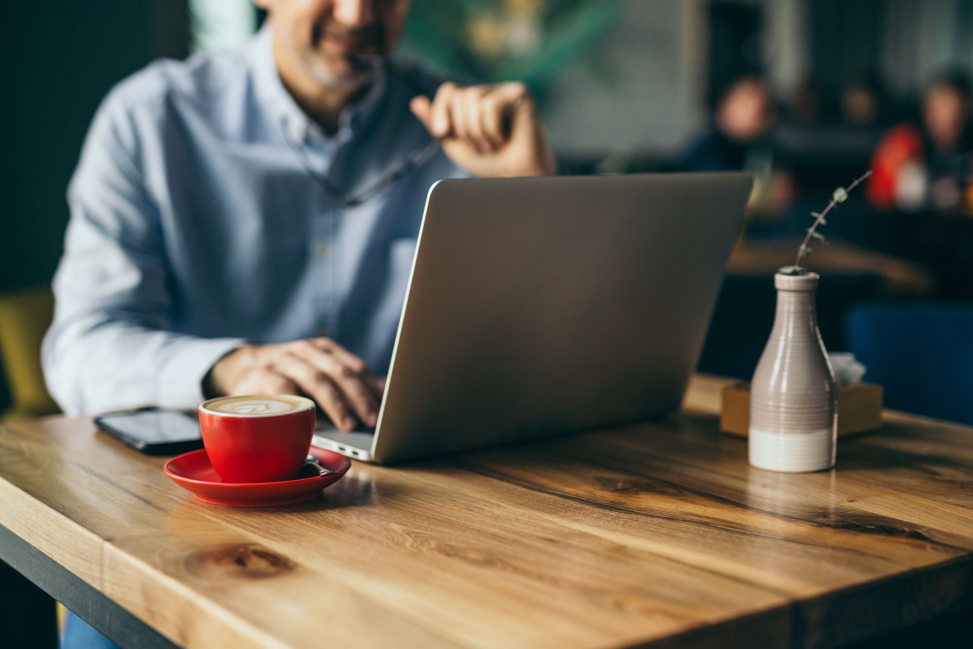 A person in a light blue shirt works on a laptop at a wooden table with a red cup of coffee and a small decorative vase.