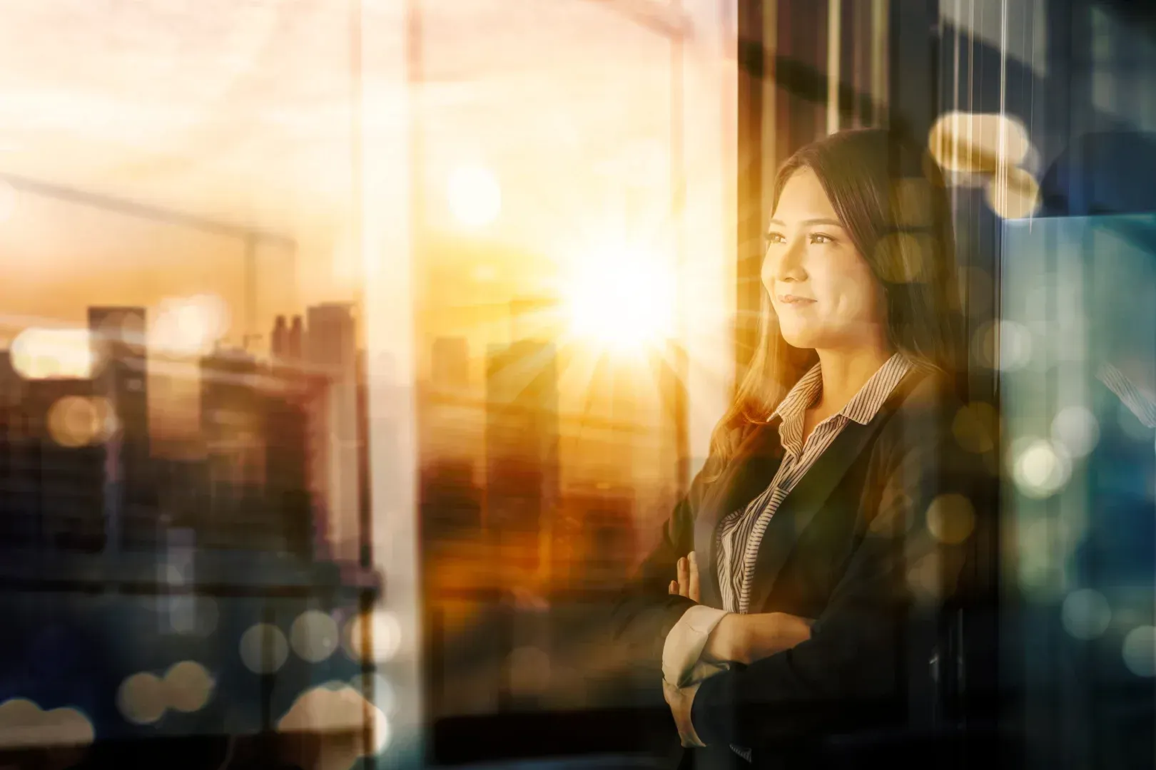Woman standing by a window at sunset, arms crossed, with warm city lights reflecting in the glass