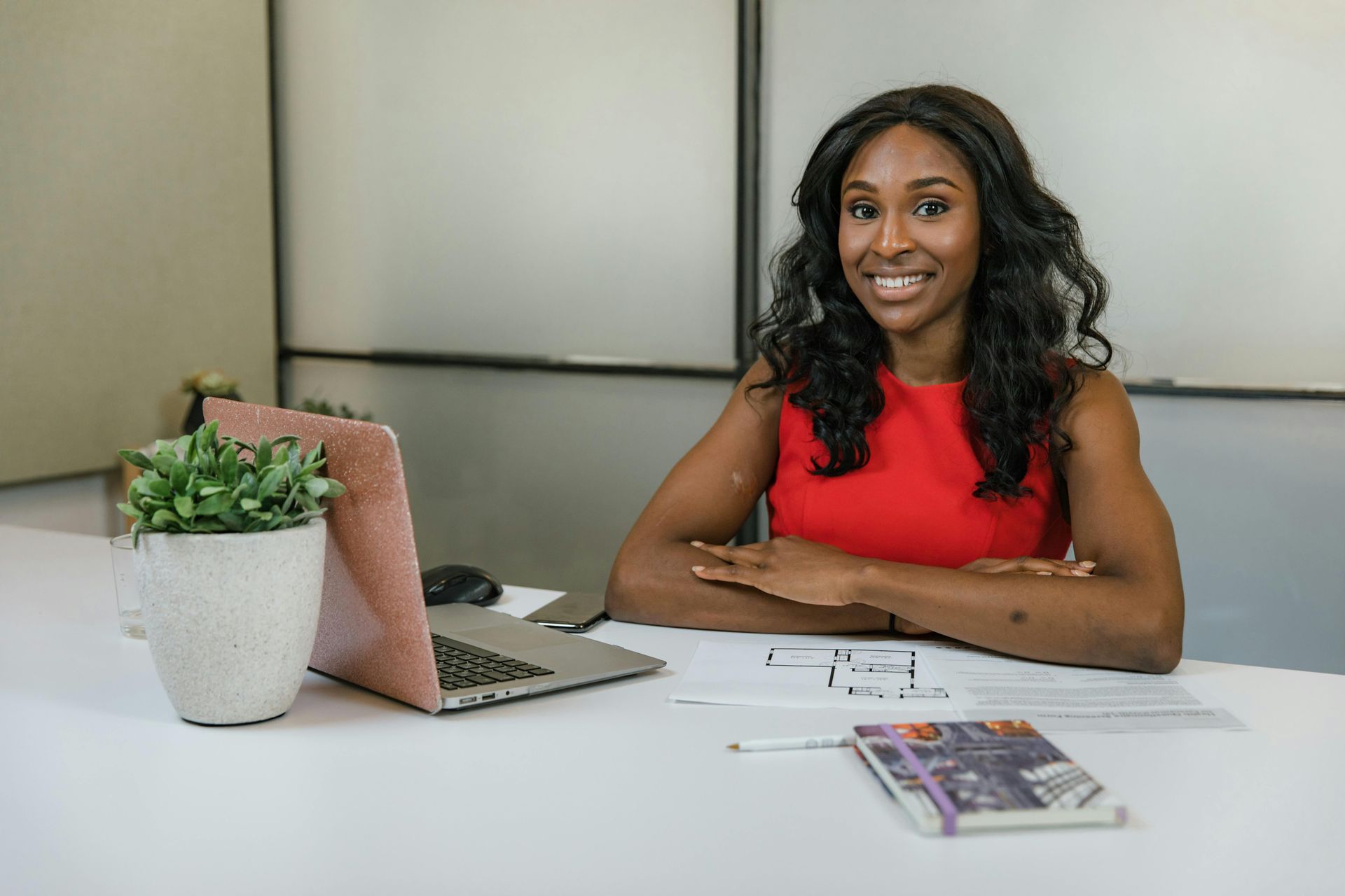 Smiling Black woman in red dress at desk with laptop and notebook.