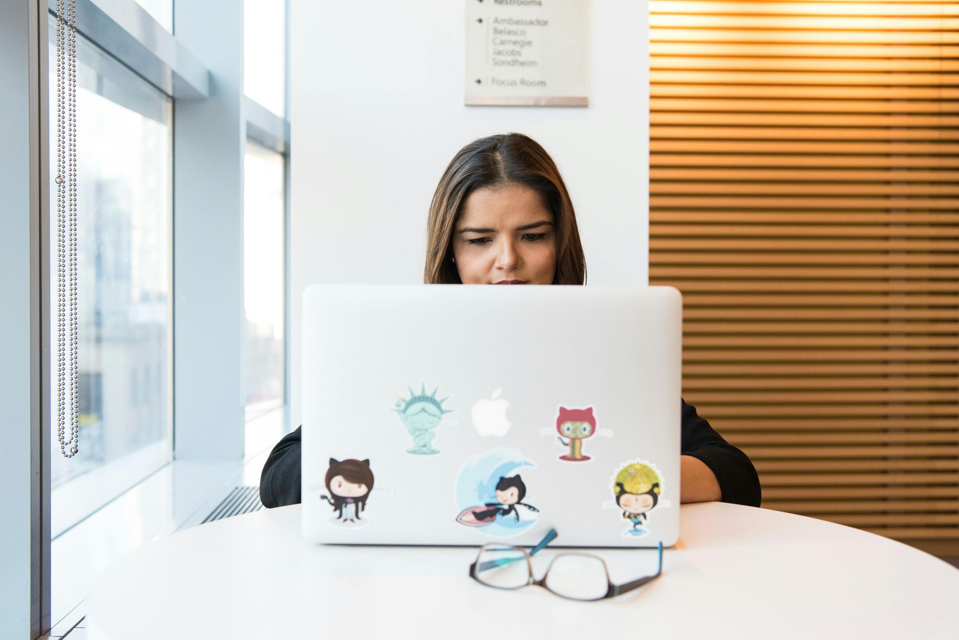 Woman using laptop at a table near a window, focused. Laptop has several stickers. Glasses on the table.