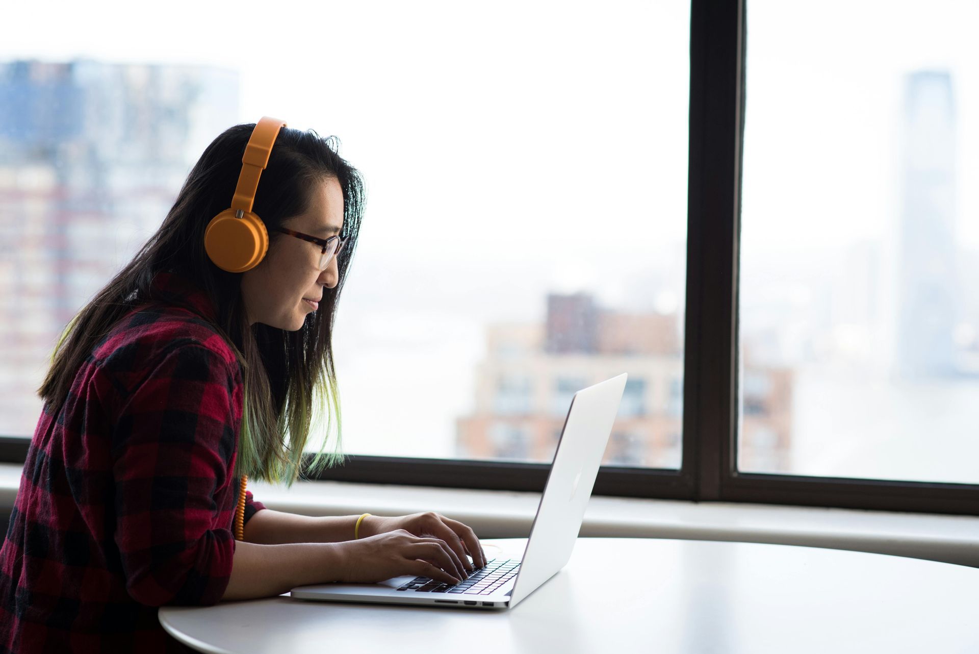 Woman with glasses and orange headphones working on a laptop by a window, overlooking a city.